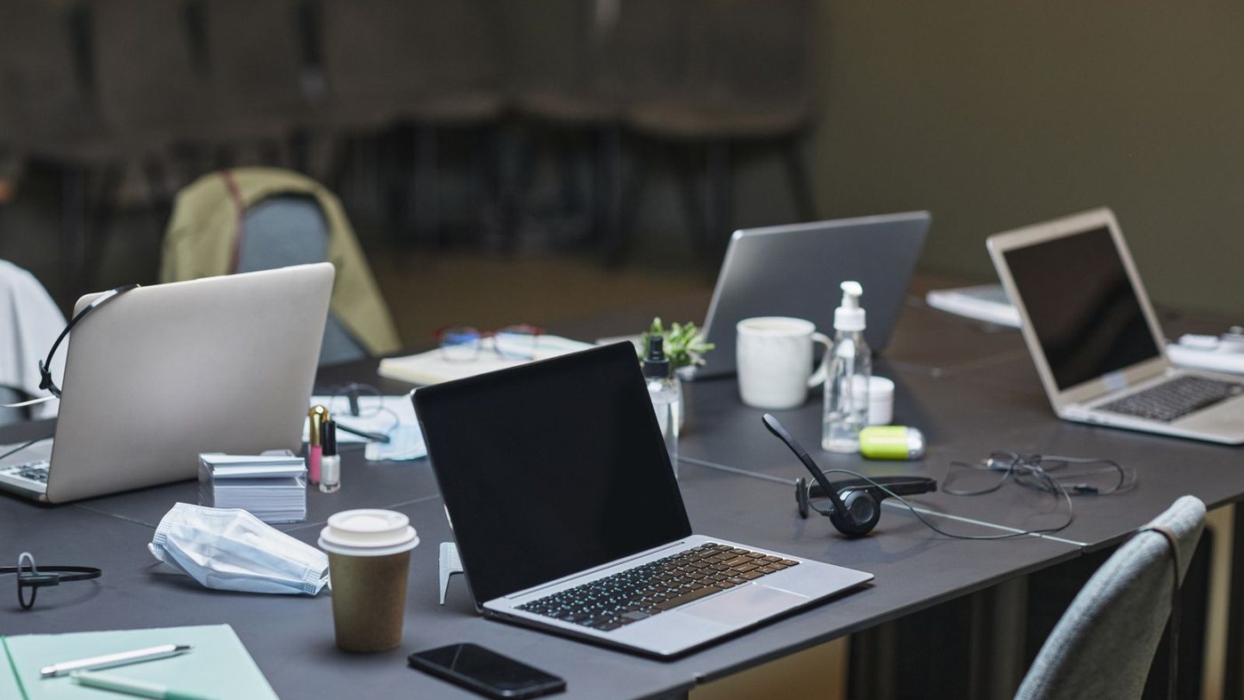 A shared worktable set up with several laptops, notebooks, headsets, and coffee