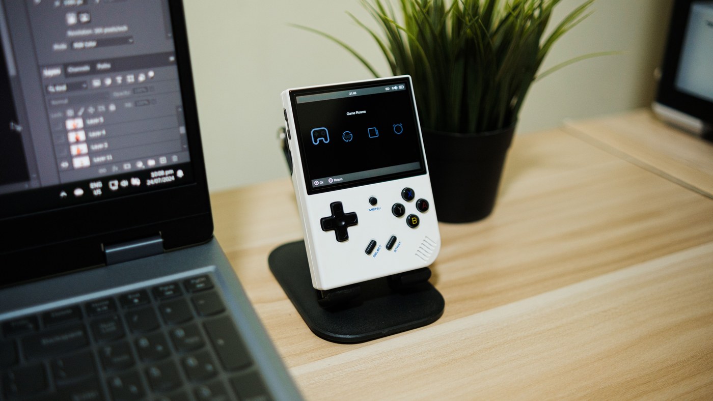 A compact retro handheld game console beside a laptop on a desk